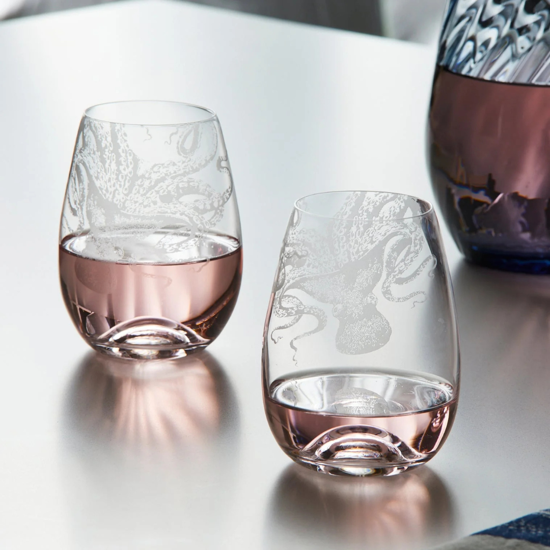 Two stemless glasses with etched octopus designs, partially filled with rosé wine, displayed on a sleek silver tabletop beside a matching glass decanter.