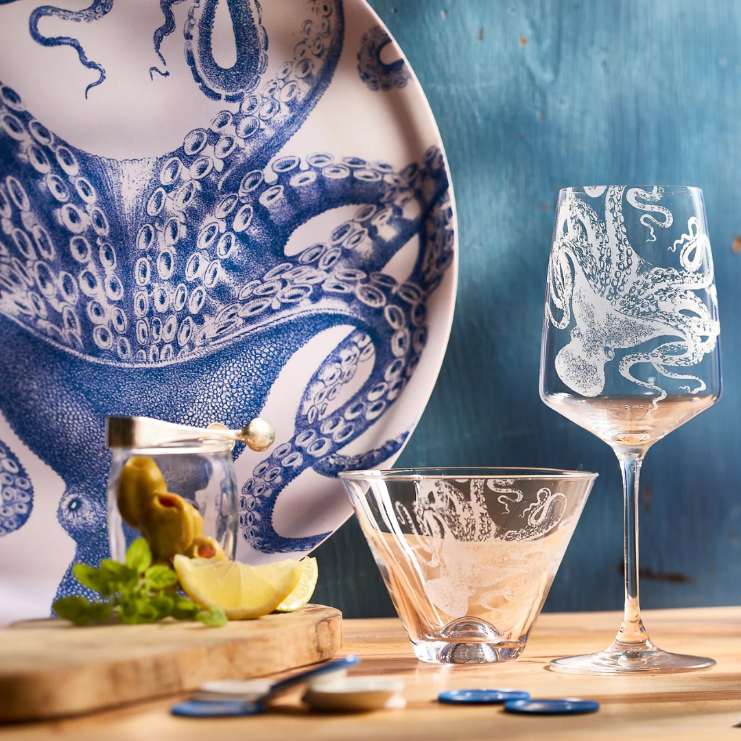 Nautical-themed glassware and dinnerware featuring intricate octopus designs in blue and white, styled with a cutting board, cocktail garnish, and sea-inspired decor against a weathered blue background.