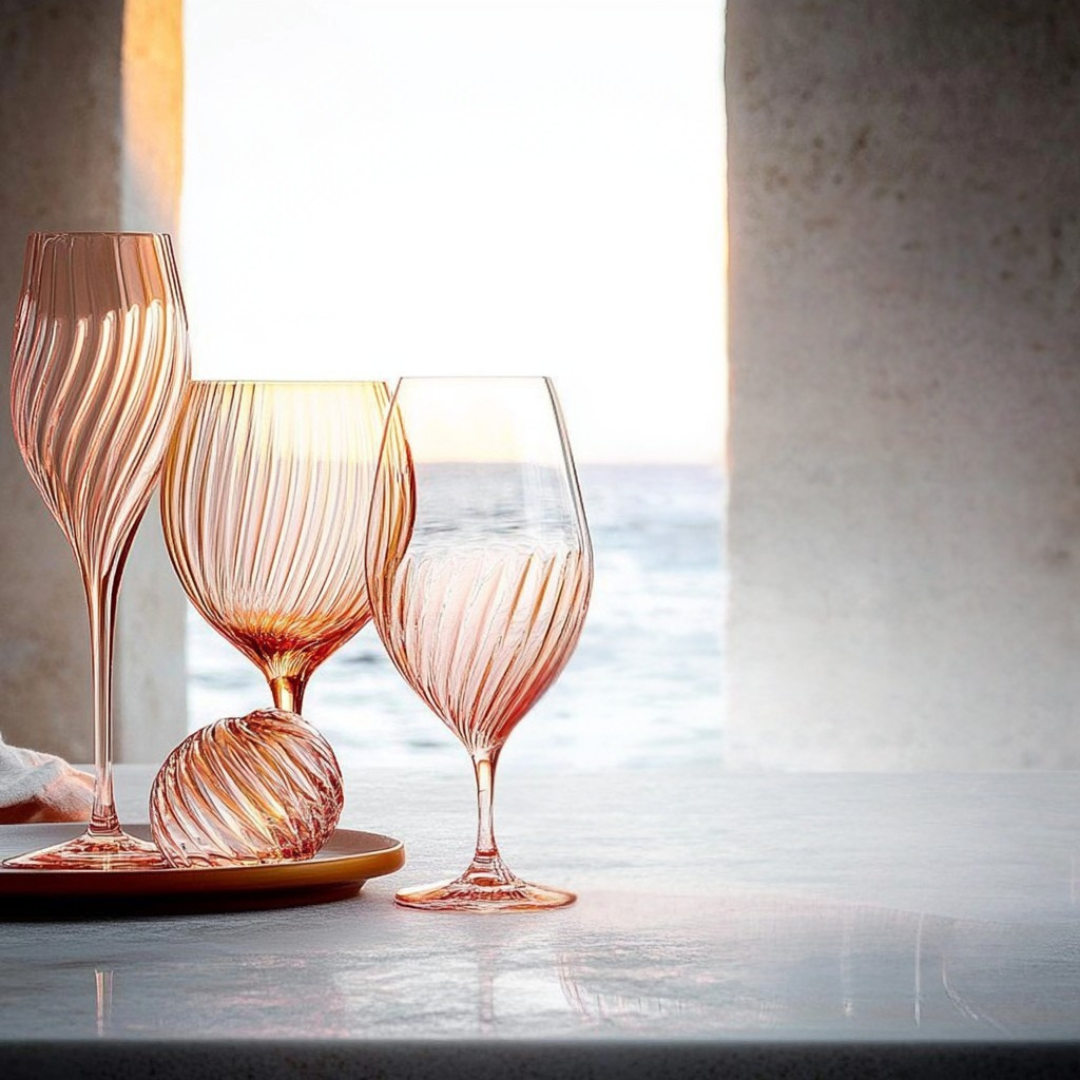Set of elegant rose-tinted ribbed glassware, including wine and champagne glasses, displayed alongside a matching decorative glass orb on a golden tray. Positioned on a stone surface, the arrangement is bathed in warm sunset light with an ocean view framed by stucco pillars, creating a luxurious coastal ambiance.
