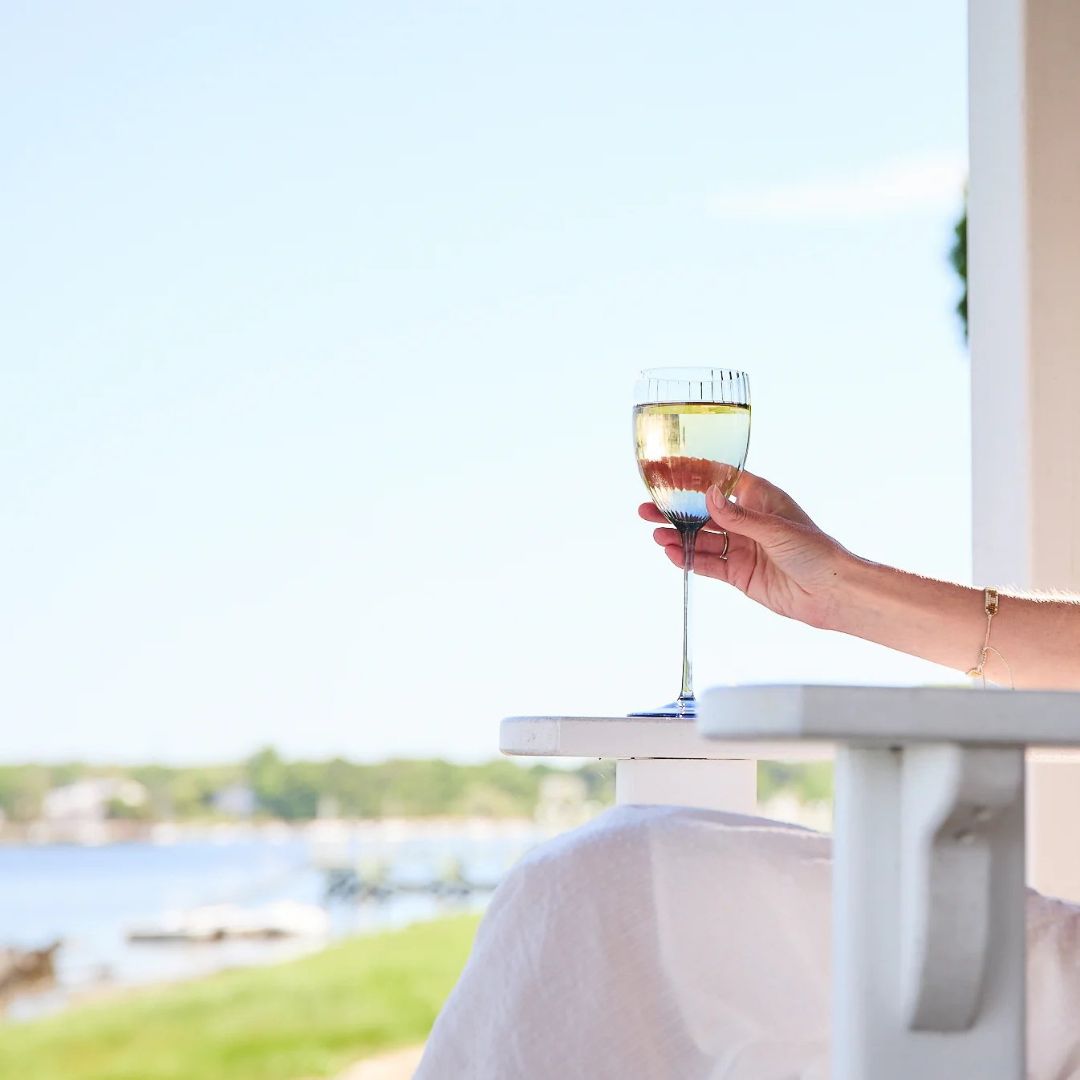 A serene coastal scene featuring a hand holding a glass of white wine, resting on the arm of a white wooden chair. The background showcases a peaceful waterfront view with blue skies, distant trees, and calm waters. The overall vibe is relaxed and luxurious, capturing the essence of a leisurely summer afternoon by the sea.