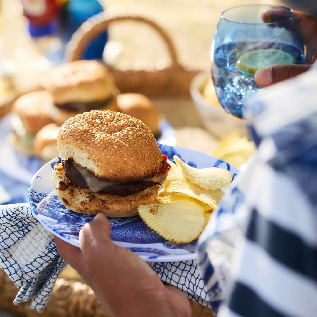 A vibrant summer picnic scene featuring a close-up of a hand holding a bright blue-tinted glass tumbler filled with sparkling water and lime slices. The glass catches the sunlight, adding a refreshing, coastal feel. In the foreground, a sesame burger and potato chips sit on a blue patterned plate, but the eye is drawn to the vivid, jewel-toned glassware that captures the relaxed energy of outdoor summer dining.