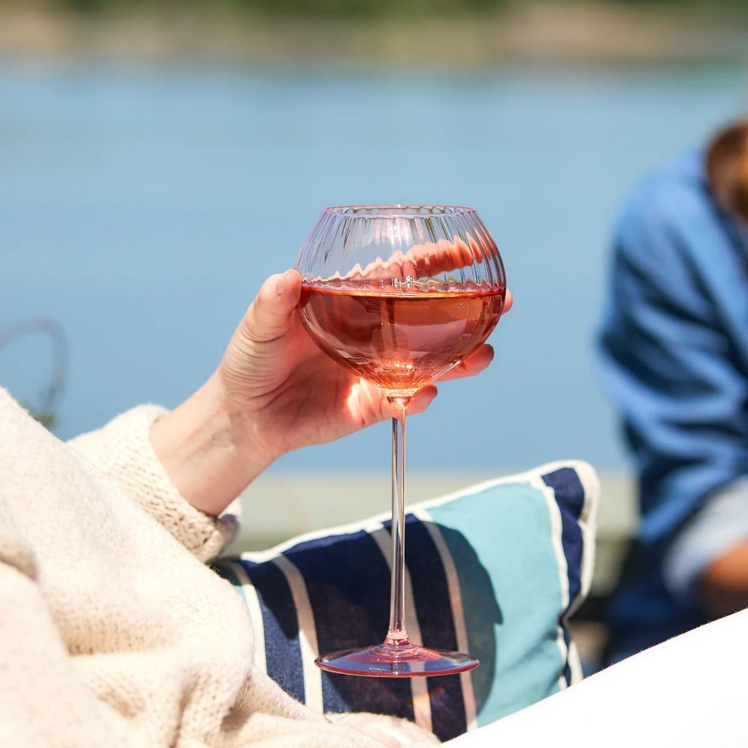 A close-up of a hand holding a round, ribbed wine glass filled with rosé, set against a serene waterfront backdrop. The person is seated comfortably on an outdoor cushion with blue and white stripes, dressed in a light sweater. The scene captures a relaxed summer moment, perfect for unwinding by the water with a chilled drink in hand.