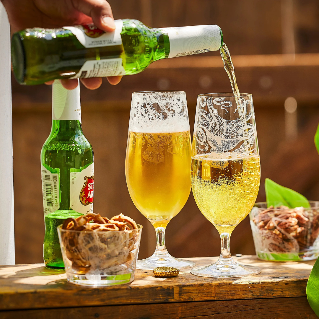Close-up of beer being poured into etched glassware on a sunny outdoor table, with two full glasses of golden beer, green Stella Artois bottles, and small bowls of pretzels and candied pecans beside them—perfect for a relaxed backyard gathering.