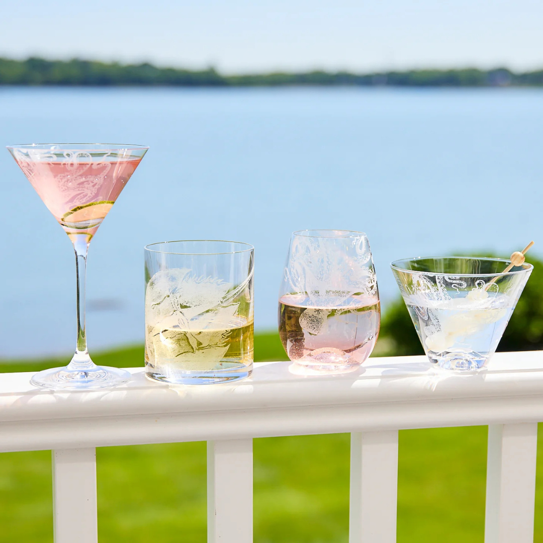 A lineup of elegant etched glassware filled with colorful cocktails, displayed on a white balcony railing overlooking a serene lakeside view on a sunny day.