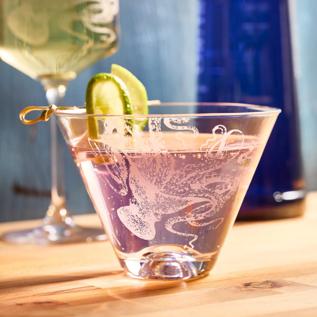 Close-up of a pink cocktail served in a clear etched martini glass featuring an octopus design, garnished with cucumber and lime slices on a skewer, with a blurred blue bottle and wine glass in the background on a light wooden surface.