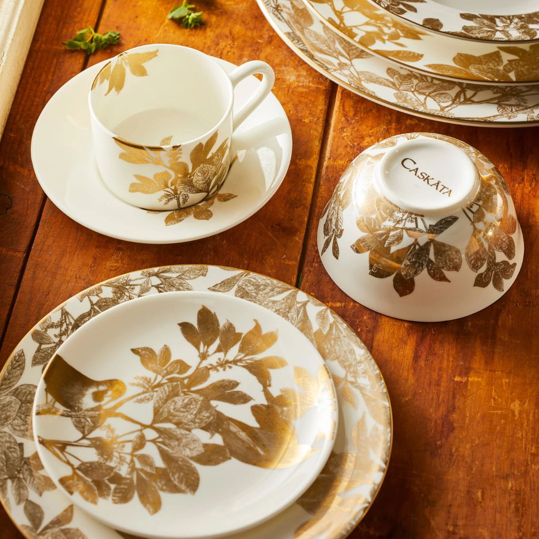 An elegant ceramic dinnerware set displayed on a rustic wooden table, featuring white plates, bowls, and a teacup adorned with intricate gold and taupe botanical illustrations. 