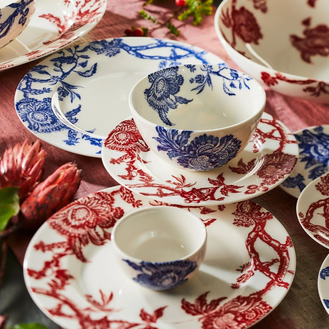 Close-up of a festive table setting featuring porcelain dinnerware with intricate red and blue floral toile patterns. The collection includes plates, bowls, and cups arranged on a rose-colored tablecloth, accented with greenery and protea flowers for a warm, seasonal feel
