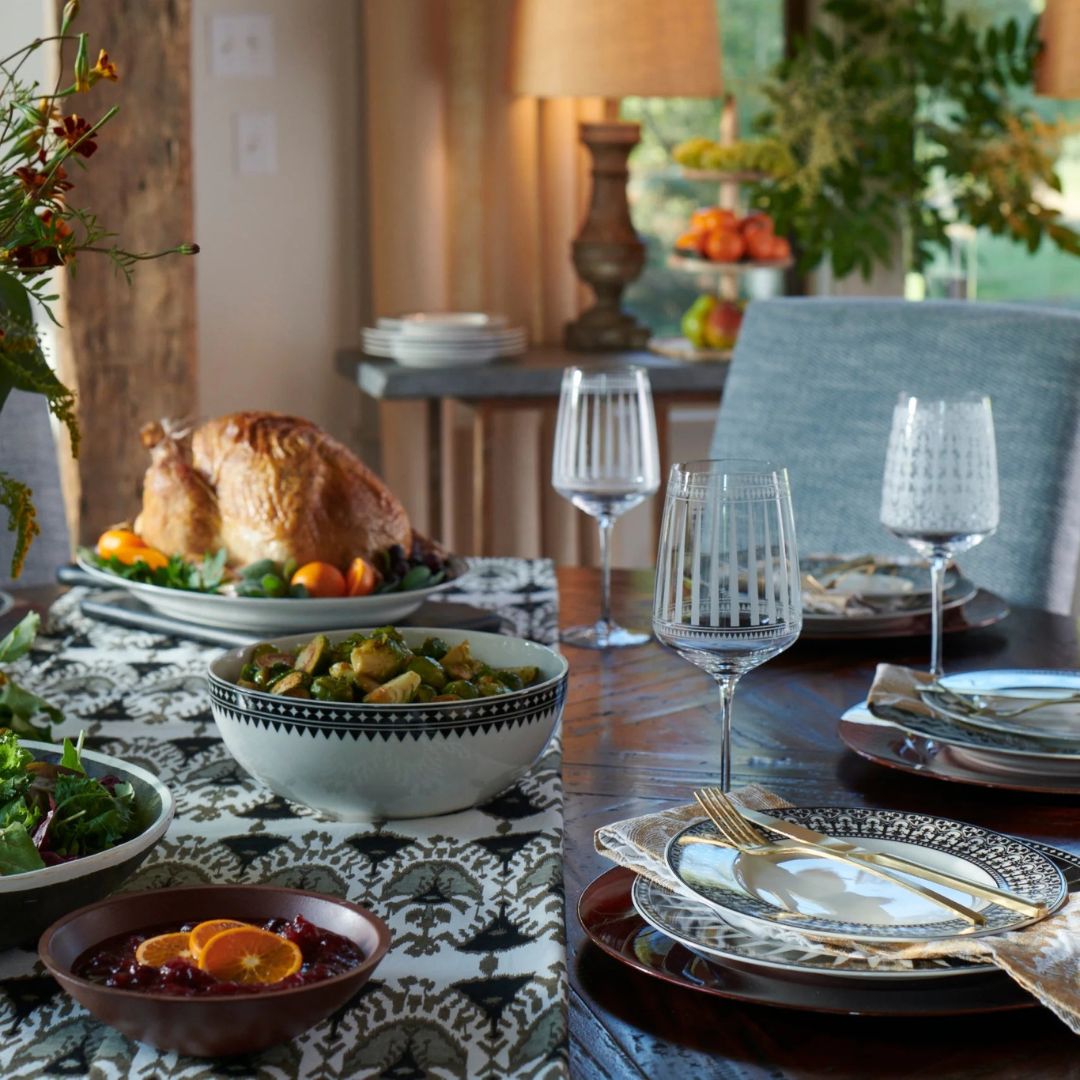 A warm and elegant dining room table set for a festive fall or holiday meal. The table features patterned dinnerware, gold flatware on folded napkins, and etched glass stemware. A roasted turkey is the centerpiece, surrounded by seasonal sides including Brussels sprouts, a mixed green salad, and cranberry sauce garnished with orange slices. The table runner adds a refined touch with its black-and-white design, while soft lighting and autumnal decor in the background create an inviting, celebratory atmosphere.