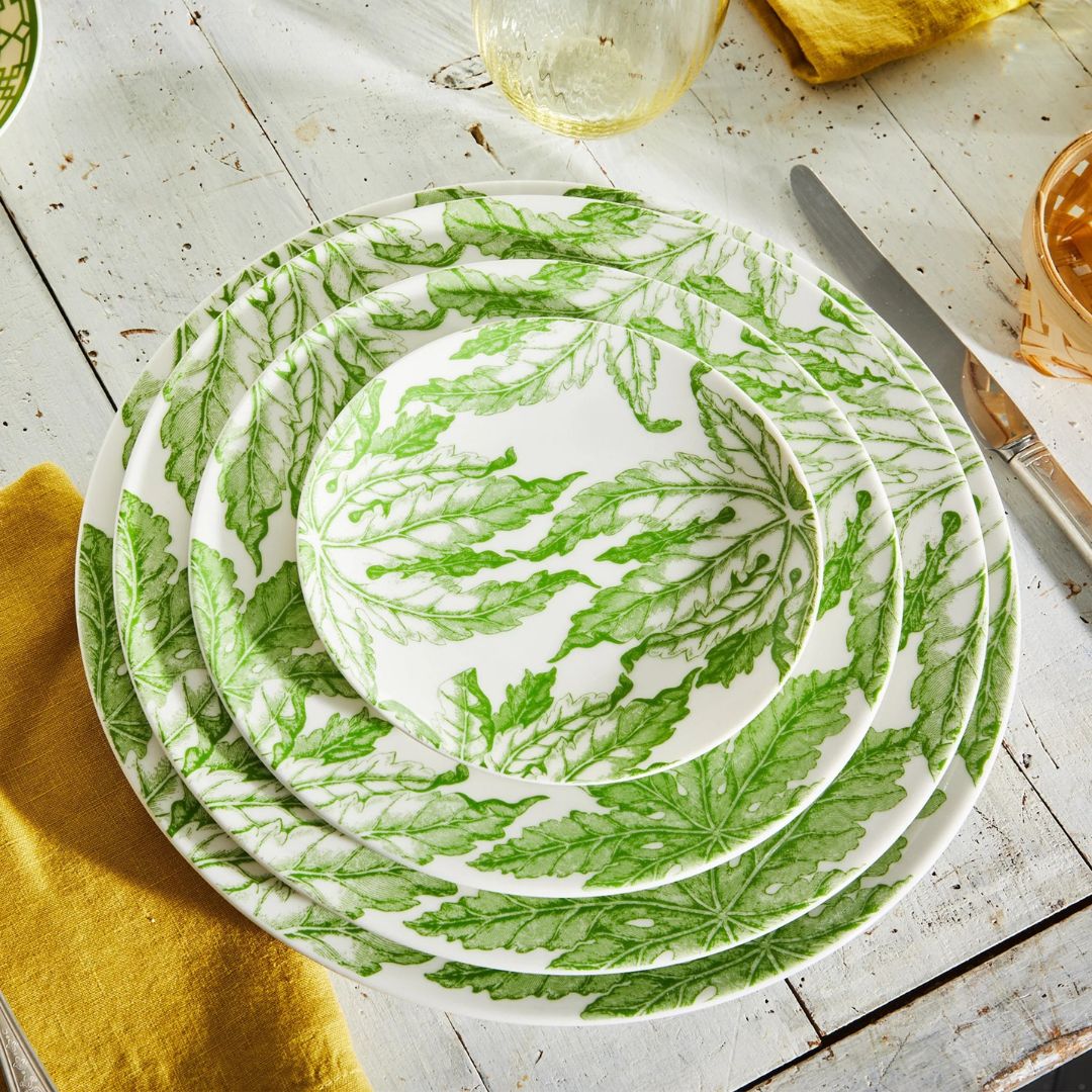 Stacked set of porcelain plates featuring a fresh green leafy pattern on a rustic white wood table. The vibrant botanical design is complemented by a yellow linen napkin, glassware, and silver cutlery, creating a cheerful, nature-inspired table setting.