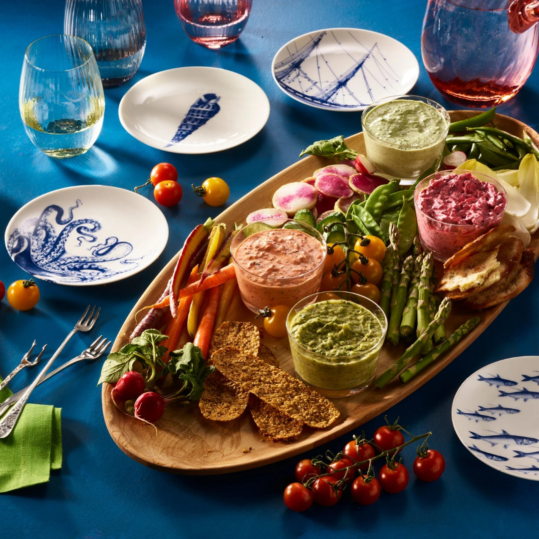 Vibrant appetizer spread on a bright blue table, featuring a large wooden platter filled with fresh vegetables, toasted bread slices, crackers, and four colorful dips in shades of green, red, and orange. Surrounding the platter are cherry tomatoes on the vine, radishes, asparagus, carrots, and snap peas. Decorative small plates with nautical blue designs and assorted glassware add a playful, coastal-inspired touch to the lively table setting.