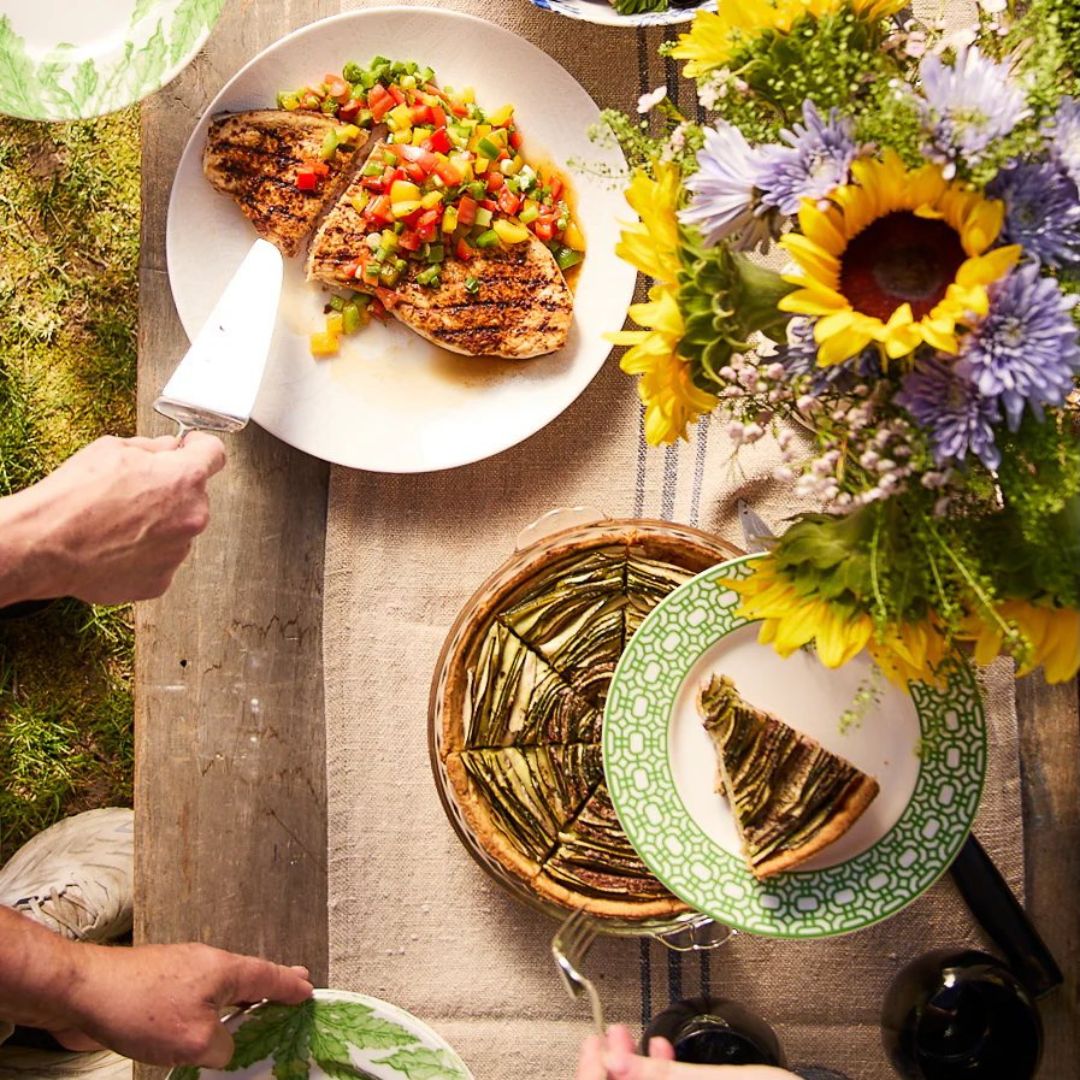 A cozy outdoor picnic-style meal set on a rustic wooden table with a natural linen runner. The spread includes a grilled chicken breast topped with colorful diced peppers, a beautifully arranged spiral vegetable tart, and a green-trimmed plate with a slice being served. Hands reach in to serve food, adding a sense of warmth and gathering. A vibrant bouquet of sunflowers, lavender, and wildflowers brightens the scene, while the surrounding grass and soft lighting evoke a relaxed, summery atmosphere perfect for sharing a homemade meal with loved ones.