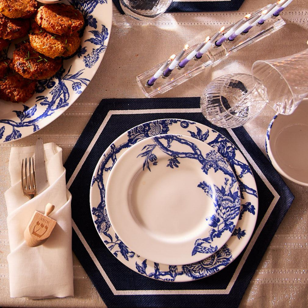 Elegant Hanukkah table setting featuring blue-and-white floral china on a navy hexagonal placemat, a white napkin wrapped around silverware with a wooden dreidel, and crystal glassware. A platter of golden latkes garnished with chives sits nearby, along with a clear menorah holding lit blue-and-white candles. The silver-toned tablecloth adds a festive, refined touch to the celebration.