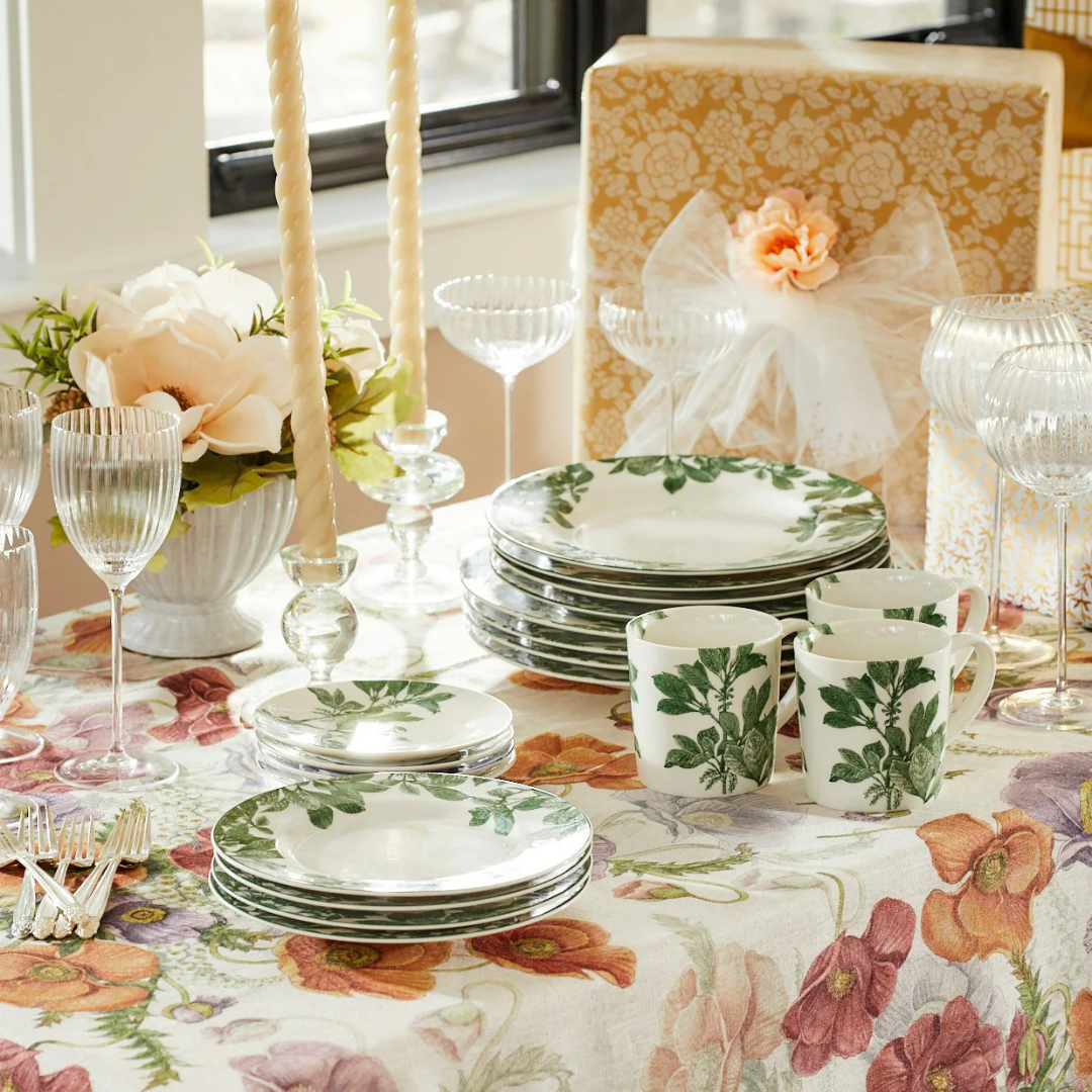 Bright and cheerful table setting featuring white dinnerware adorned with green botanical prints, neatly stacked alongside matching mugs. The table is covered with a floral-patterned tablecloth in soft spring hues, complemented by ribbed glassware, twisted ivory taper candles in glass holders, and a white floral centerpiece. In the background, elegantly wrapped gifts with floral bows add a celebratory touch, creating a fresh and festive atmosphere perfect for spring gatherings.