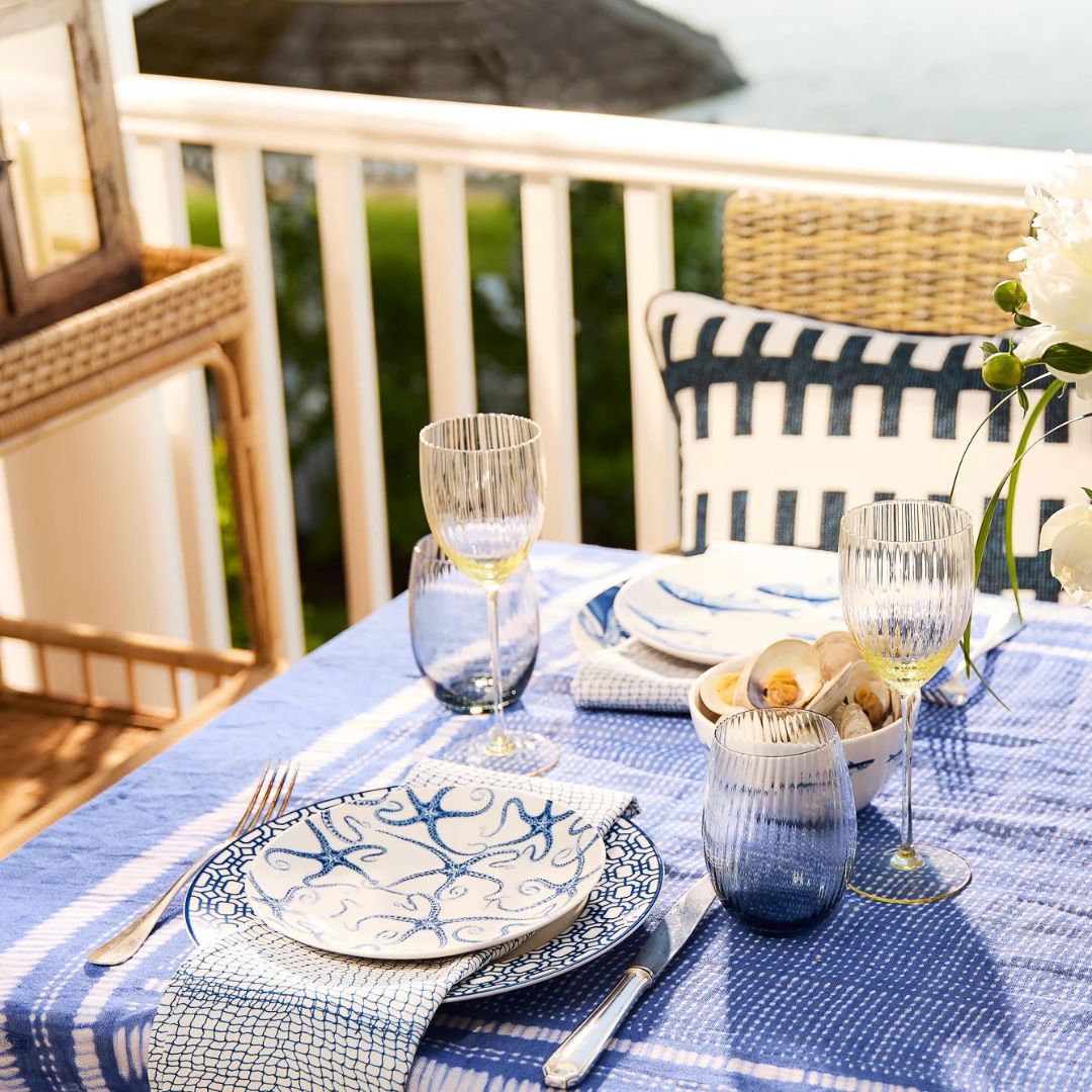 A breezy coastal dining setup on a balcony overlooking the water, featuring a blue and white theme. The table is dressed with a blue plaid tablecloth and set with patterned blue-and-white plates, textured napkins, and ribbed glassware with a hint of gold. A bowl of clams adds a nautical touch, while a bold blue-and-white cushion on a wicker chair enhances the relaxed, seaside ambiance. Perfect for an elegant summer lunch or dinner by the sea.