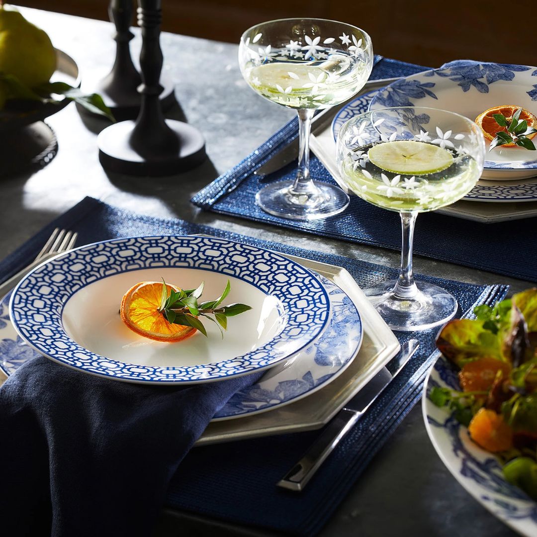 Elegant tablescape featuring blue and white patterned dinnerware, navy placemats, and etched coupe glasses with floral designs. A dried orange slice with greenery sits at the center of each plate, adding a fresh, seasonal touch. The setting is accented with a navy napkin and surrounded by moody lighting and a vibrant mixed greens salad.