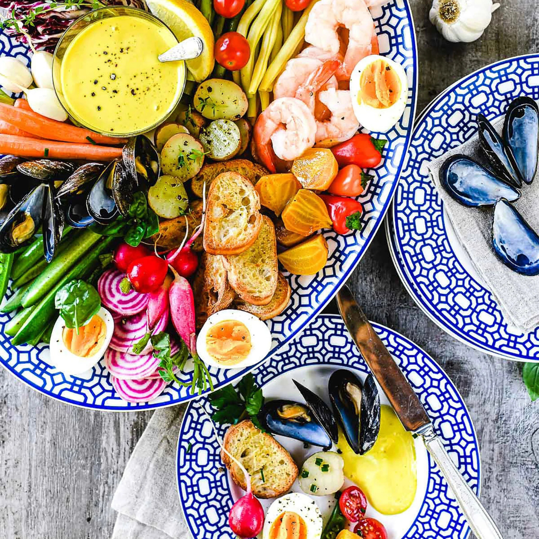 Vibrant seafood and vegetable platter arranged on a blue and white patterned plate, featuring mussels, shrimp, hard-boiled eggs, roasted beets, radishes, green beans, tomatoes, crostini, and a bowl of golden dipping sauce. A smaller matching plate is served with a selection of the same colorful ingredients.