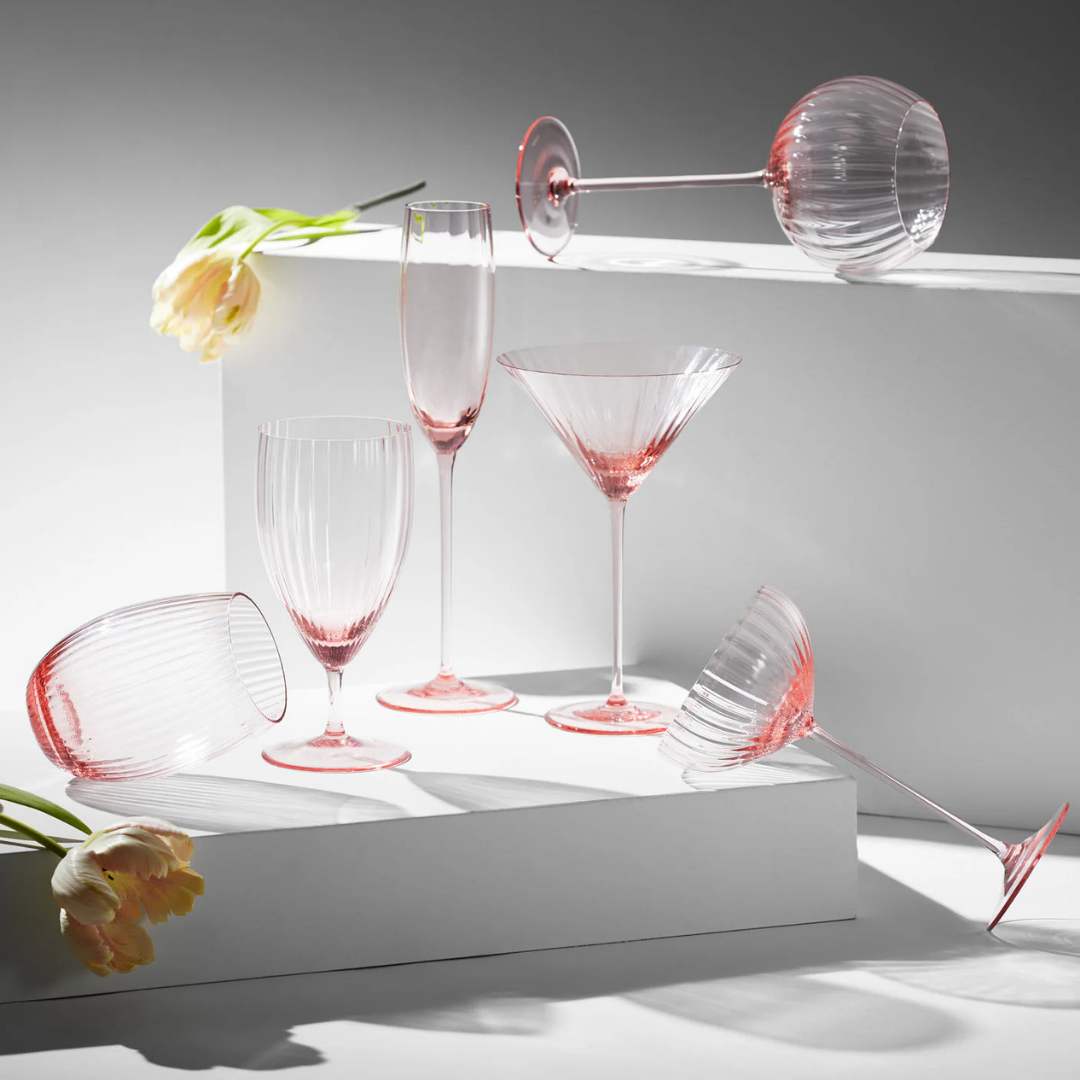 Stylized product display of elegant ribbed glassware in soft blush pink tones, featuring a variety of shapes including wine glasses, champagne flutes, martini glasses, and goblets. The glasses are arranged both upright and tilted across minimalist white platforms, accented by delicate yellow tulips. Dramatic lighting and shadows enhance the modern, romantic aesthetic of the composition.